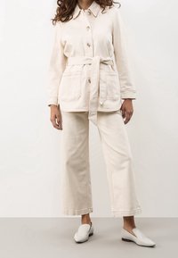 Off-white denimjacka med knyt i midjan, två framfickor, ihop med matchande vida byxor och slip-on-skor. Slät textur genomgående.