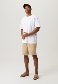 Biała bawełniana koszulka, beżowe szorty i brązowe klapki. Prosty krój z luźnym dopasowaniem. Brak widocznych wzorów lub zdobień.