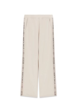 Beżowe spodnie dresowe z kieszenią z przodu i bocznymi paskami z napisem „H Sails Guess”, noszone z krótką beżową kurtką i wzorzystym granatowym dołem bikini.