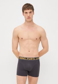 Dunkelgraue Boxer-Shorts mit einem gelben Logo-Bund, die ein figurbetontes Design und eine glatte Textur aufweisen, getragen von einem Modell ohne Shirt.