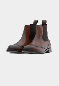 Bottes Chelsea en cuir marron avec côtés élastiques noirs, détails brogués sur la pointe et languettes pour un enfilage facile. Semelle plate en caoutchouc.