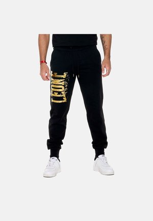 Homme portant un pantalon de jogging noir avec un grand texte doré usé sur la jambe gauche et des baskets blanches sur un fond uni.