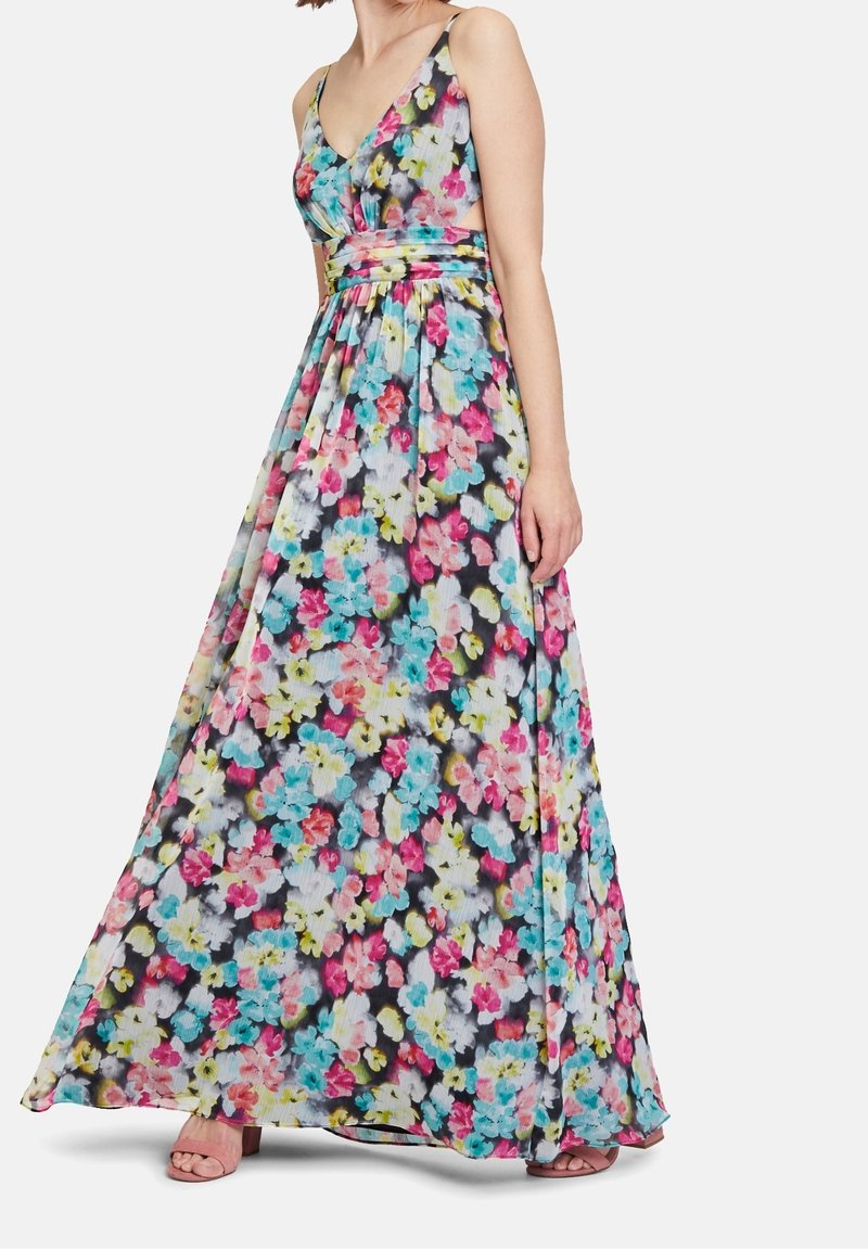 Femme portant une robe longue sans manches à fleurs, cintrée à la taille avec des découpes sur les côtés, debout avec une main détendue le long du corps.