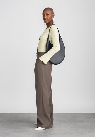 Melnas ādas plecu soma ar noapaļotu formu. Modelis valkā gaiši zaļu ribotu topu, brūnas pielāgotas bikses un baltas smailes kurpes.