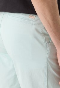 Pantalon bleu clair avec poche passepoilée arrière et écusson logo BOSS sur la ceinture, porté avec un top noir visible.