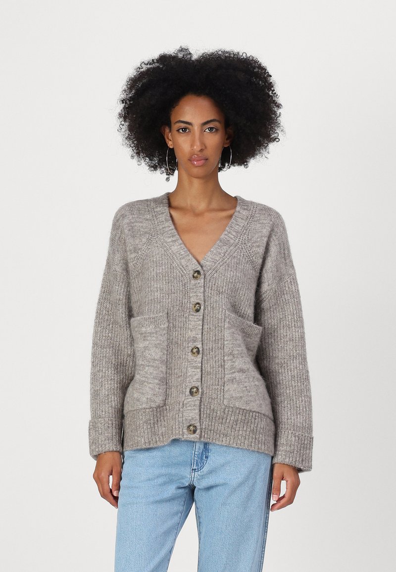 Pelēks adīts kardigāns ar V-veida kakla izgriezumu, ribotām piedurknēm un divām priekšējām kabatām. Iespējas ietver lielos pogas un mīkstu, strukturētu audumu.