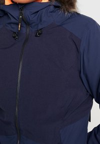 Giacca impermeabile blu navy con corpo testurizzato, fodera arancione e cappucci con pelliccia. Presenta una chiusura a zip e cordini regolabili.