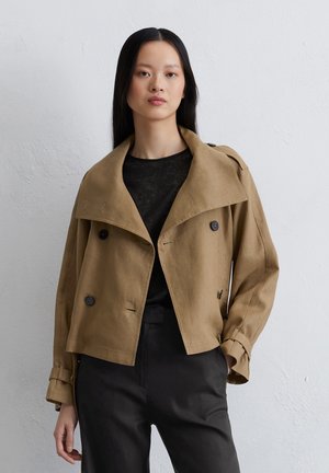Tan cropped jacket with a wide collar, double buttons, and rolled cuffs, paired with a black shirt and tailored black pants.