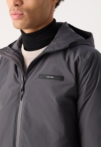 Giacca grigia con cappuccio realizzata in materiale sintetico liscio, con zip frontale e una piccola patch nera con il logo Calvin Klein sul petto.