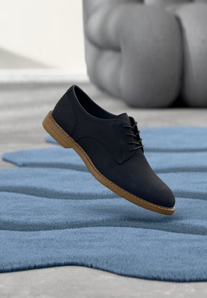 Chaussure en cuir noire à lacets avec semelle marron suspendue au-dessus d'un tapis bleu texturé, avec un canapé gris flou en arrière-plan.