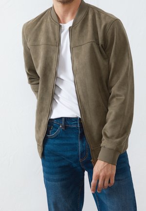 Homme portant une veste bomber zippée vert olive par-dessus un t-shirt blanc et un jean bleu, debout devant un fond uni.