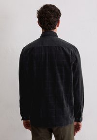 Czarna koszula ze sztruksu o fakturowanej powierzchni, z subtelnymi wzorami w kratę, zapinanymi mankietami i ostrym kołnierzem, widziana od tyłu.