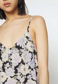 Top de alças preto com padrão floral, apresentando grandes flores amarelas e roxas, um decote em V, alças ajustáveis e três botões pretos decorativos.
