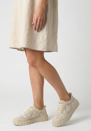 Beine einer Person, die ein beiges Leinenkleid und cremefarbene, spitzenmusterartige Plateausneaker trägt, stehend vor einem schlichten hellen Hintergrund.