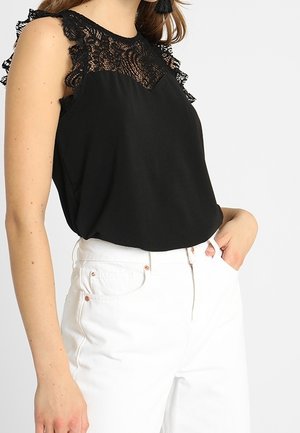 Vrouw draagt een zwart mouwloos kanten topje gecombineerd met witte, hooggetailleerde jeans, staat met één hand in de zak tegen een witte achtergrond.