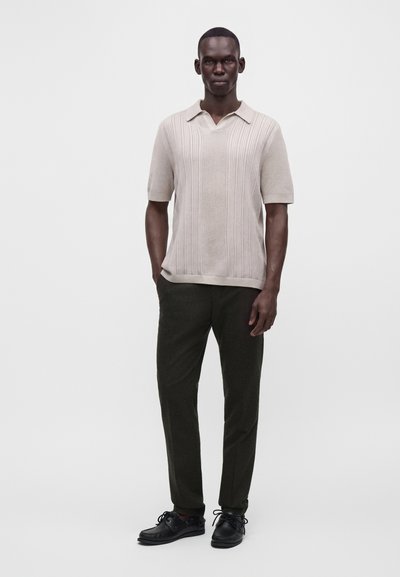 Man wearing a light beige short-sleeve knit polo shirt, dark trousers, and black lace-up shoes, standing with hands in pockets.