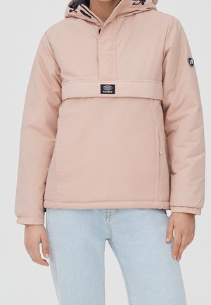 Person trägt eine hellrosa Kapuzen-Winterjacke mit einer Fronttasche und hellblauen Jeans vor einem einfarbigen Hintergrund.