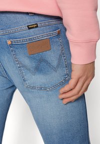 Jasnoniebieskie dżinsy Wrangler z brązową skórzaną naszywką na tylnej kieszeni, z charakterystycznym wzorem zszycia w zygzak oraz dopasowanym pomarańczowym szyciem.