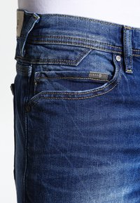 Tmavě modré džíny z denimu s designem na pět kapes, viditelným prošíváním a značkovou visačkou na přední kapse. Mají klasický střih a texturu.