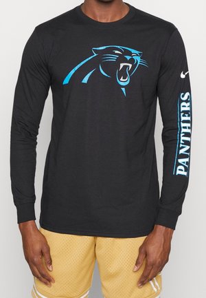 Homme portant un t-shirt noir à manches longues avec un logo de panthère rugissante sur la poitrine et le texte « PANTHERS » sur la manche droite, assorti à un short jaune.