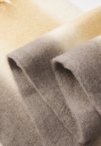 Villane tekk, millel on gradienttriibud beežis, kreemis ja hallis. Pehme, tekstuuriga kanga sees on üksteisega kattuvad osad. Sobib sooja ja mugavuse tagamiseks.