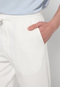 Witte trainingsbroek met een elastische tailleband en zijzakken, gemaakt van zachte stof. Een hand ligt casual in de zak, wat een ontspannen pasvorm benadrukt.