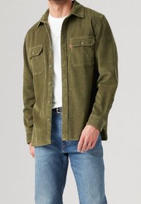 Camicia in corduroy verde oliva con due tasche sul petto, chiusura a bottoni e colletto classico. Indossata con una t-shirt bianca e jeans in denim.