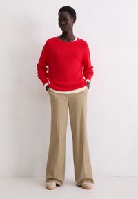 Rode ribgebreide trui met een crèmekleurige halslijn, gecombineerd met beige wijde broekspijpen en lichtgekleurde schoenen. Staand voor een neutrale achtergrond.