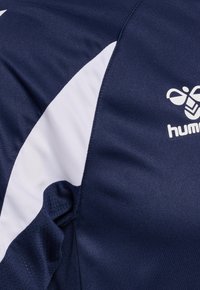 Maillot de sport bleu marine avec une bande blanche incurvée et le logo hummel sur la poitrine, montrant la texture du tissu et les détails des coutures.