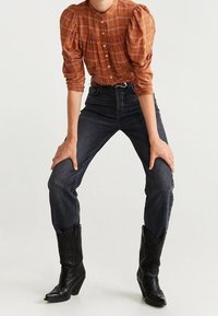 Blouse à carreaux couleur rouille avec des manches bouffantes, associée à un jean foncé taille haute et des bottes de cow-boy noires à bouts pointus et à motif texturé.