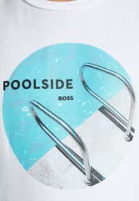 Bílý tričko s kulatým grafikou na téma tyrkysového bazénu, betonového okraje a stříbrného bazénového žebříku. Text zní "POOLSIDE BOSS."