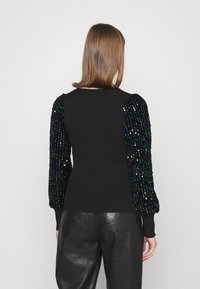 Pull noir côtelé avec des manches volumineuses ornées de sequins aux teintes vertes, bleues et violettes. Porté avec un pantalon en cuir, mettant en valeur une silhouette ajustée.