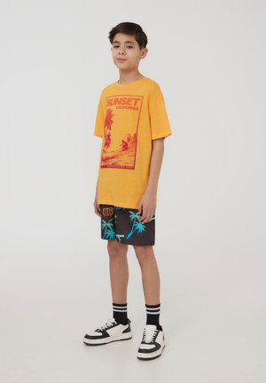 Maglietta gialla a maniche corte con stampa grafica rossa. Pantaloni corti neri con motivo di palme blu. Sneakers bianche e nere, calzini a righe nere.