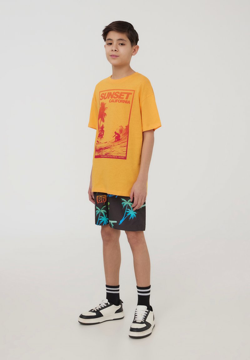 Maglietta gialla a maniche corte con stampa grafica rossa. Pantaloni corti neri con motivo di palme blu. Sneakers bianche e nere, calzini a righe nere.