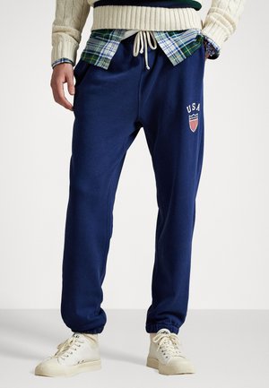 Marinblå sweatpants med elastisk midjeband och dragsko. USA-logotyp på vänster lår. Bärs över en rutig skjorta, ihop med vita sneakers.