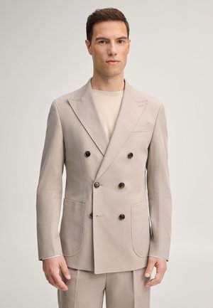 Man staand tegen een egale achtergrond, gekleed in een lichtbeige dubbelrijig colbert en bijpassende broek met een neutraal overhemd.