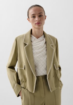 Blazer corto color verde oliva con cuciture bianche a contrasto, due tasche frontali e revers piegati; abbinato a un top leggero e texturizzato.