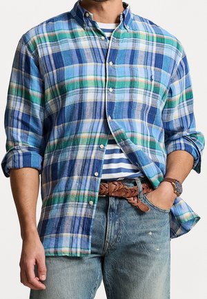 Hombre con camisa de cuadros azul y verde abotonada sobre una camiseta de rayas azul y blanca, con vaqueros azules, cinturón trenzado marrón y reloj de pulsera.
