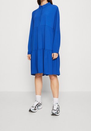 Femme portant une robe bleue ample à manches longues avec un col et une jupe à volants, des chaussettes blanches et des chaussures de sport blanches avec des touches noires et métalliques.
