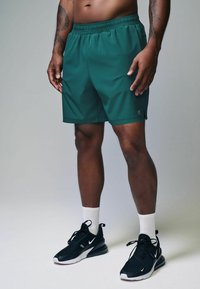Mørkegrønne atletiske shorts med elastisk talje og sidelommer, parret med sorte sneakers og hvide ankelsokker.