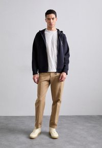 Hanorac cu fermoar de culoare navy peste un tricou crem cu model, asortat cu pantaloni khaki și adidași de culoare deschisă. Design minimalist, texturi moi.