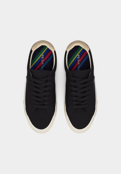 Paire de baskets basses noires avec semelles blanches, contreforts beiges au talon, lacets noirs, et semelles intérieures rayées colorées avec texte de la marque.