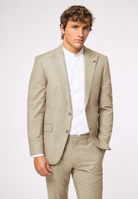 Helles beige Anzug mit strukturierter Webart, einreihiger Jacke und geraden Hosen, kombiniert mit einem weißen Button-Down-Hemd.