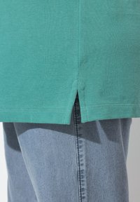 T-shirt in cotone turchese con spacchi laterali all'orlo, abbinato a jeans di denim azzurro, che mostra un tessuto testurizzato e una vestibilità casual.