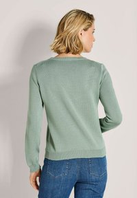 Frau mit kurzen blonden Haaren, die einen hellgrünen Pullover und eine blaue Jeans trägt, steht mit dem Rücken zur Kamera vor einem schlichten Hintergrund.