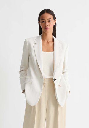 Vrouw met lang donker haar die een witte blazer, een wit topje en beige wijde broek draagt, staand tegen een effen lichte achtergrond.