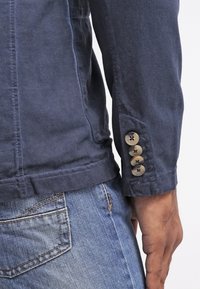 Primo piano del braccio di una persona che indossa una giacca blu navy con quattro bottoni sul polsino e jeans di denim blu con una tasca posteriore.