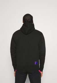 Sudadera con capucha negra, de mangas largas, con puños acanalados y un pequeño parche de logo colorido en el dobladillo. Textura de tela suave.