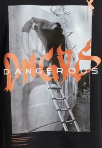 T-shirt en coton avec un fond noir, une impression photographique grise, et un texte orange en gras affichant "DANGEREUX" dans une police stylisée.