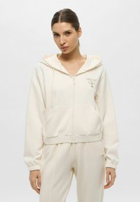 Felpa con zip di colore crema, dotata di cordino regolabile, tasche laterali e polsini a coste; presenta un logo ricamato sul petto. Abbinata a pantaloni coordinati.